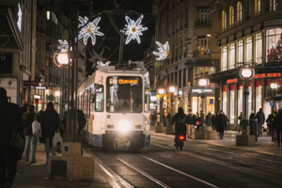 Genebra à noite: rua central iluminada, com bonde, luzes suspensas e fachadas antigas.